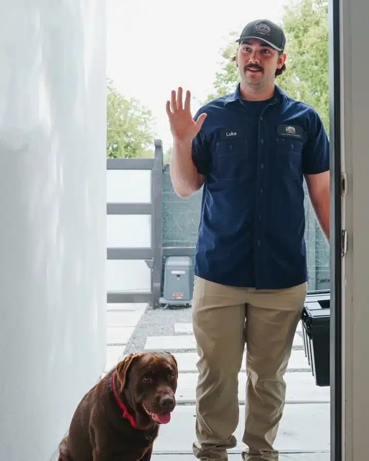 Luke's plumber waving at the front door of a Houston home, with the company's Labrador by his side